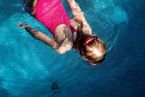 Yläpuolella on pieni söpö lapsi tyttö urheilu vaaleanpunainen uimapuku ja suojalasit sukellus vedenalainen kirkas sininen vesi sisäuima-allas pimeässä illalla. Lapset vesiurheilu terveellinen toiminta - Valokuva, kuva