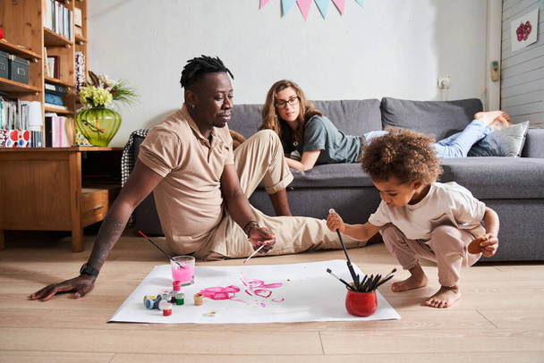 Man looking with interest at his little curly daughter sitting at the floor and drawing - Fotoğraf, Görsel
