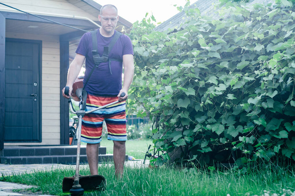 Człowiek w goglach, szortach i T-shirtach kosi trawnik z przycinarką do benzyny na tle winnicy i wejścia do domu - Zdjęcie, obraz