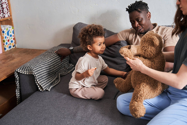 Curly girl looking at her lovely teddy bear while her mother holding him - Fotografie, Obrázek