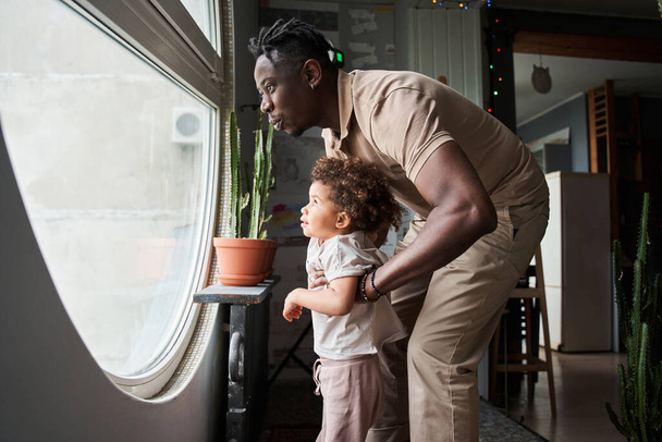 Multiracial man looking at the big round window with his little cute daughter - Photo, image