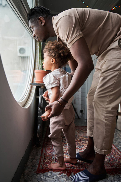 Multiracial man looking at the big round window with his little cute daughter - Photo, Image