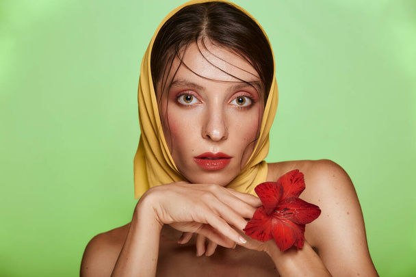 Ładna kobieta z czerwonym kwiatem w dłoniach przy twarzy. Jej głowa w modnej chustce nagie ramiona jasny model skóry z różowym makijażem. Koncepcja pielęgnacji skóry i spa na zielonym tle. - Zdjęcie, obraz