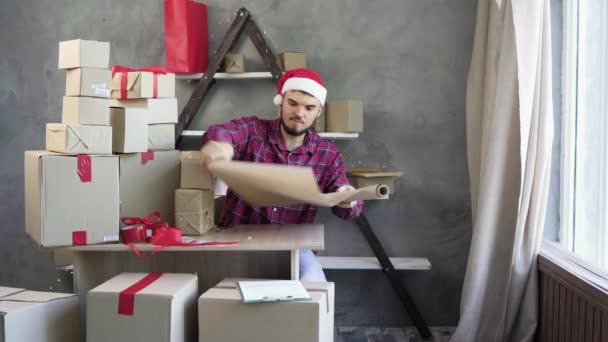 Vendita di Natale e piccole imprese, l'uomo caucasico siede a tavola nel suo ufficio a casa e prepara le merci per il mandrino. prende un po 'di carta da imballaggio. - Filmati, video