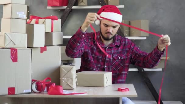 Běloch, malý podnikatel, sedí v domácí kanceláři v klobouku Santa Clause a přivazuje červenou mašli na krabici. koncept vánočního prodeje. - Záběry, video