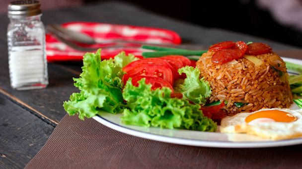 Gebratener Reis mit chinesischer Wurst serviert in einem weißen Teller mit Spiegelei, Tomaten und Salat. - Foto, Bild