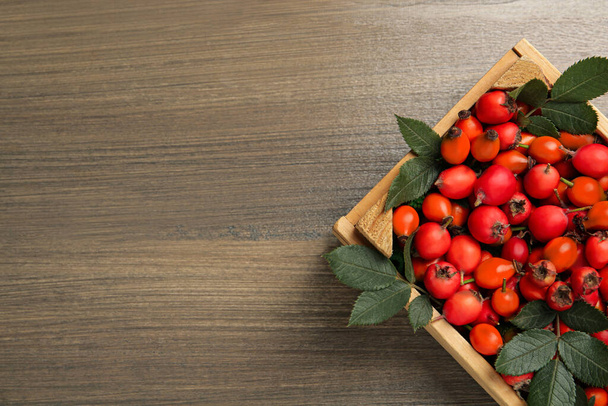 Ripe rose hip berries with green leaves in crate on wooden table, top view. Space for text - Foto, immagini