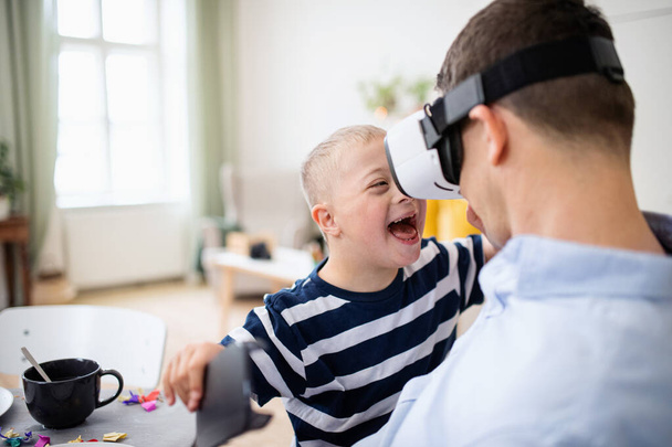 Father with happy down syndrome son indoors at home, using vr goggles. - Фото, изображение