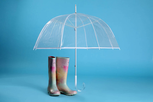 Open transparent umbrella and rubber boots on light blue background - Zdjęcie, obraz