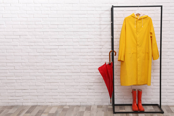 Elegante estante con paraguas, impermeable y botas de goma cerca de la pared de ladrillo blanco. Espacio para texto - Foto, Imagen