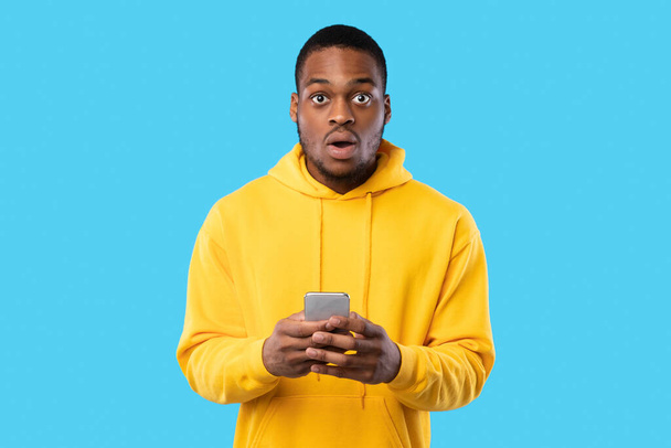 Megdöbbent afro-amerikai Guy holding telefon pózol át kék háttér - Fotó, kép