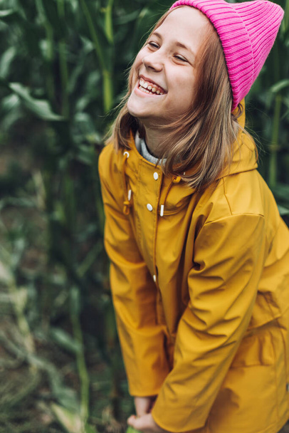 Stijlvol tiener meisje in gele regenjas en hete roze hoed lachen op maïsveld - Foto, afbeelding
