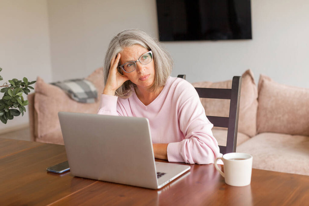 Pensive middle-aged woman sit at table in living room using laptop look in distance thinking or pondering, thoughtful senior gray-haired female distracted lost in thoughts feel lonely or sad at home - Foto, Imagen