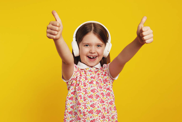 Little cute girl listening music in white headphones and showing thumbs up - Fotó, kép