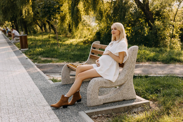 Zwangere vrouw in witte jurk zittend op een bankje in een park op een zonnige dag. - Foto, afbeelding