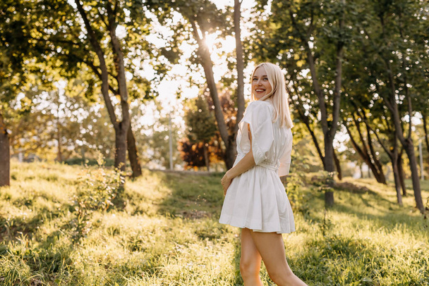 Ciężarna kobieta spacerująca w parku w słoneczny dzień, uśmiechnięta, ubrana w białą sukienkę. - Zdjęcie, obraz