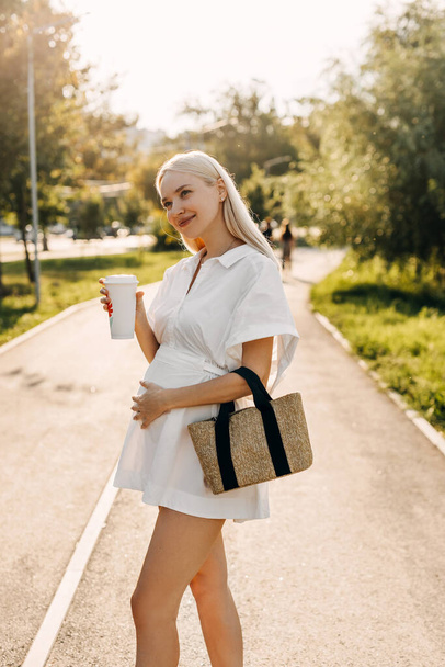 Terhes nő sétál a parkban, kezében egy to go kávéscsésze, mosolygós. - Fotó, kép