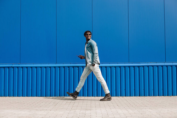 african guy walks with a phone around the city, against the background of a blue building, makes a purchase online through the phone, a full-length portrait, steps to the side - Φωτογραφία, εικόνα