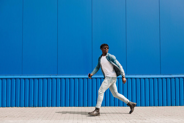 african guy walks with a phone around the city, against the background of a blue building, makes a purchase online through the phone, a full-length portrait, steps to the side - Fotó, kép