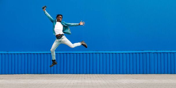 african guy jumping with a phone in his hands, in the city against the background of a blue building, concept of success - 写真・画像