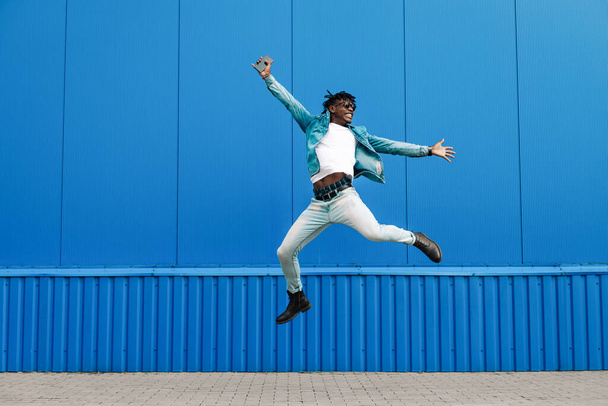 african guy jumping with a phone in his hands, in the city against the background of a blue building, concept of success - Photo, image