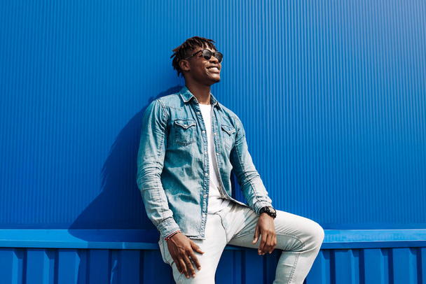 african man in sunglasses stands against a blue wall of a building in the city, handsome black man smiling - Foto, Bild