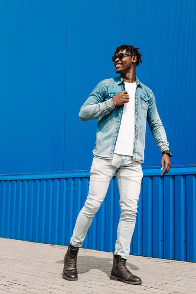 fashion african man in sunglasses in trendy modern clothes posing against the background of a blue building, on the street - Fotó, kép