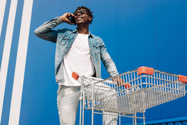 african black man walking with a shopping cart talking on the phone, making a phone call, shopping online talking with an operator, against background of a blue building - Photo, image