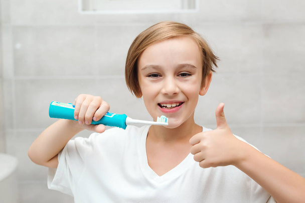 A mosolygós fiú törődik a fogai egészségével. Gyerek fogat mos elektromos kefével a fürdőszobában. Fogászati higiénia minden nap. Egészségügy, gyermek- és fogápolás. Boldog fiú fogmosás. - Fotó, kép