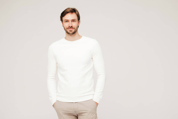 Portrait of handsome smiling hipster lumbersexual businessman model wearing casual white sweater and trousers. Fashion stylish man posing against grey background in studio.Looking at camera - 写真・画像