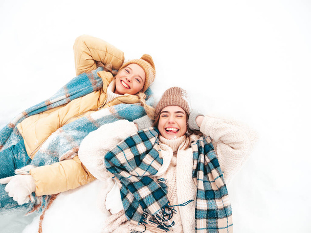 Две молодые красивые улыбчивые женщины-хипстеры в трикотажных теплых одеждах и шарфах позируют на улице в парке. Положительные чистые модели, лежащие в снегу. Наслаждаюсь зимними моментами. Концепция Рождества - Фото, изображение