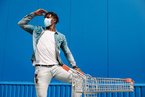 afro american black man walking with a shopping cart in a protective mask from coronavirus, looking far into the distance - Foto, Imagen