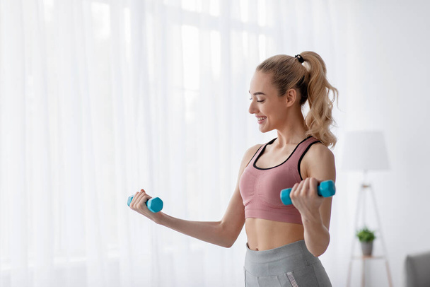Cheerful european cute young blonde woman lifts up dumbbells at home, work out alone, takes care of body - Photo, Image