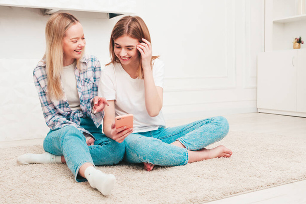 Two young beautiful smiling women. Sexy carefree models sitting in white interior in apartments or hotel room. Female looking at smartphone screen. They using cellphone apps indoors - Foto, immagini