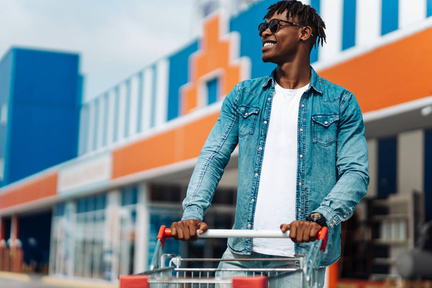 afro american black man with shopping trolley in front of the mall, shopping in the mall, successful shopping mall, wearing goggles in sun - Foto, imagen
