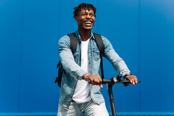 young african american man on an electric scooter, rides on the road, in city against the background of a blue building, rushes to school or work - Foto, immagini