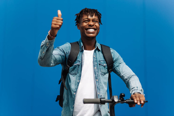 young african american man on an electric scooter, rides on the road, in the city against the background of a blue building, shows a gesture: thumbs up, super, class, joy - Foto, imagen