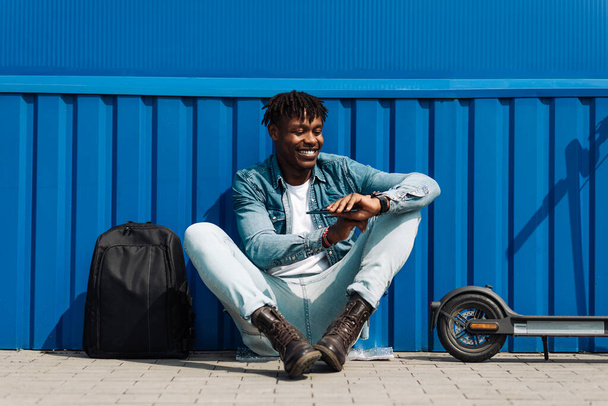 An African American man with an electric scooter and a phone in his hands sits against a blue wall, answers an SMS message through the clock, via the Internet 5g, online correspondence - Fotó, kép
