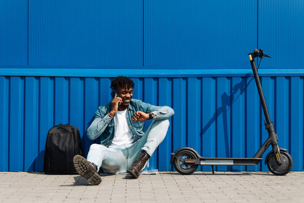 An African American man with an electric scooter and a phone in his hands sits against a blue wall, answers an SMS message through the clock, via the Internet 5g, online correspondence - Фото, изображение