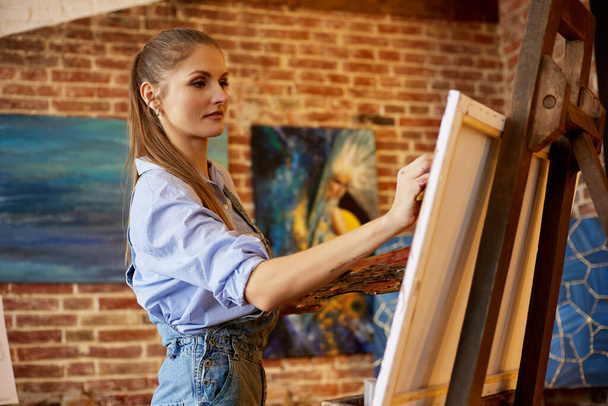 Young beautiful female artist painting on canvas using oil paintings and art brush. Painter creating artwork in art studio. Relaxation, leisure, hobby, stress management - Valokuva, kuva