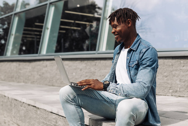 black African American typing on a laptop on the street in front of a glass building, sitting online, working on the road, high-speed mobile internet, 5g, 4g - Photo, Image