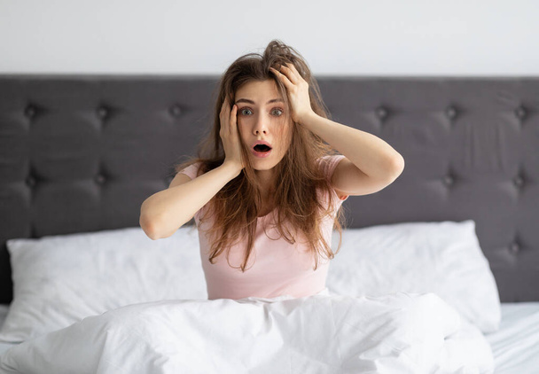 Geschockte Millennial-Frau, die zu Hause auf dem Bett sitzt, den Kopf vor Schreck hält, zu spät zur Arbeit kommt - Foto, Bild