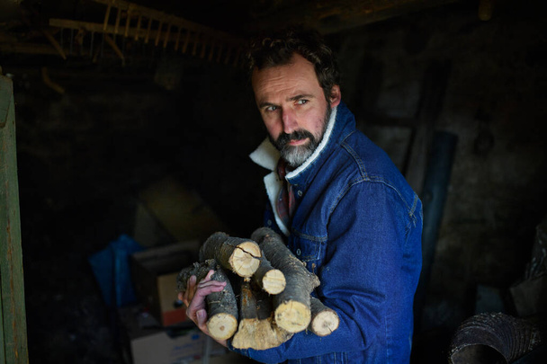 Portrait of poor mature man holding firewood at home, looking at camera, poverty concept. - Fotografie, Obrázek
