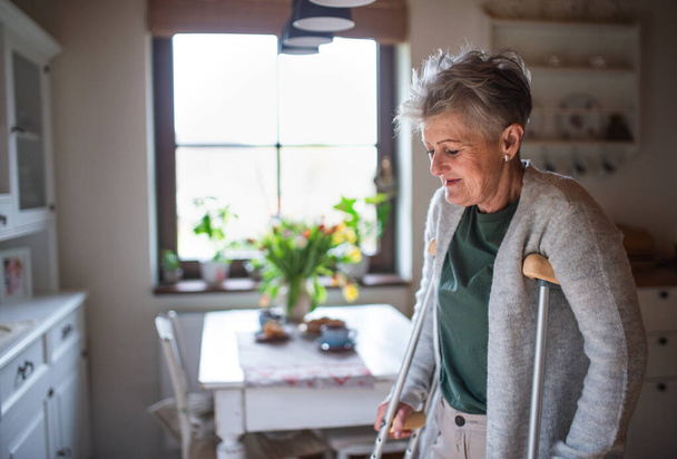 Senior woman with crutches indoors at home, walking in kitchen. - Photo, image