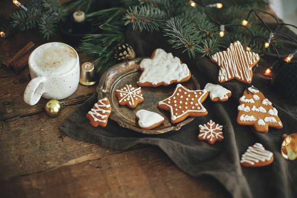 Χριστουγεννιάτικα μπισκότα μελόψωμο, καφέ σε κομψό λευκό κύπελλο, κλαδιά ελάτης, ζεστά φώτα σε χαρτοπετσέτα και ρουστίκ ξύλινο τραπέζι. Ωραία κυκλοθυμική εικόνα. Χειμώνας. Καλές γιορτές.! - Φωτογραφία, εικόνα