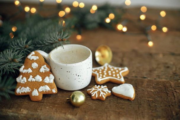 Kerst peperkoek koekjes, koffiekop, sparren takken en bauble op de achtergrond van warme lichten bokeh op rustieke houten tafel. Moody atmosferisch beeld. Winterlandschap hygge. Seizoenen begroeting - Foto, afbeelding