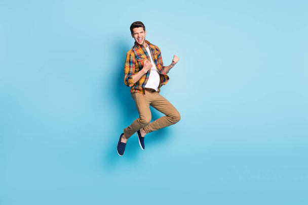 Photo of carefree lucky young gentleman wear checkered shirt jumping high rising fist smiling isolated blue color background - Foto, afbeelding