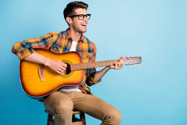 Photo of pretty adorable guy dressed plaid shirt spectacles smiling siting chair playing guitar empty space isolated blue color background - Φωτογραφία, εικόνα
