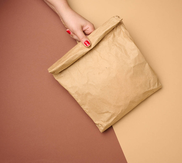 mano sosteniendo una bolsa de papel llena de papel kraft marrón sobre fondo marrón, de cerca - Foto, imagen