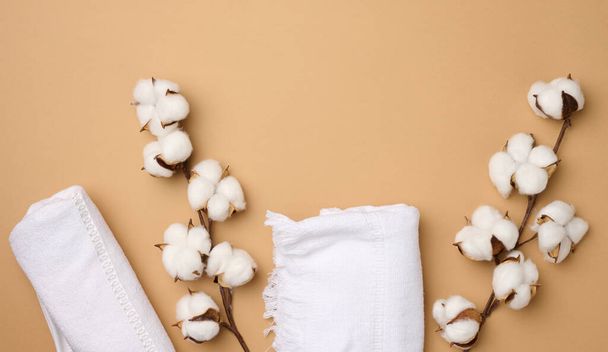 toalha de algodão branco dobrado terry e raminhos de flor de algodão em um fundo marrom claro, vista superior - Foto, Imagem